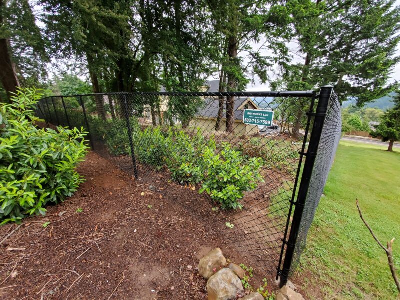 Black chain-link fence running along a landscaped bed of shrubs with a Big Works LLC sign attached.
