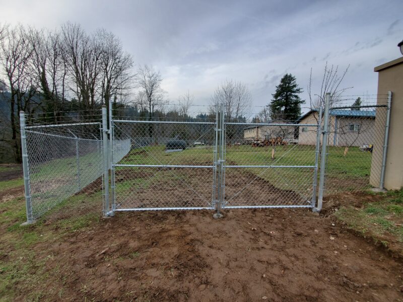 Galvanized chain-link double drive gate and fence line on a rural lot with bare graded dirt in the foreground.
