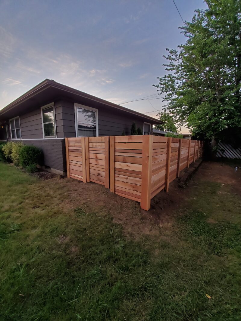 Horizontal-slat cedar privacy fence wrapping the corner of a gray ranch house at dusk