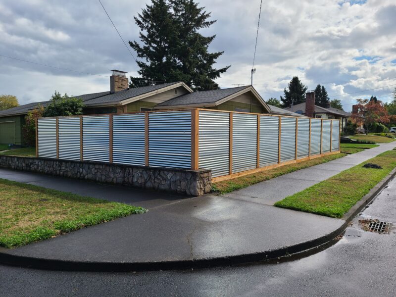 Corrugated metal privacy fence with cedar frames built atop a stone retaining wall on a corner lot.