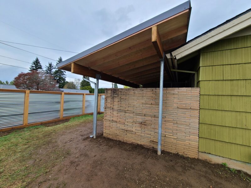 Slanted wood-framed patio cover on steel posts with a stacked-lath privacy wall and corrugated metal fence.