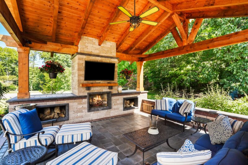 Timber-frame covered outdoor living room with stone fireplace, mounted TV, ceiling fan, and cushioned lounge seating on a slate patio.