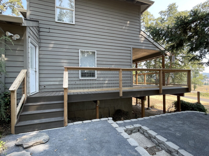 Composite deck with cedar-framed hog-wire railing and stairs wrapping the back of a gray two-story home.