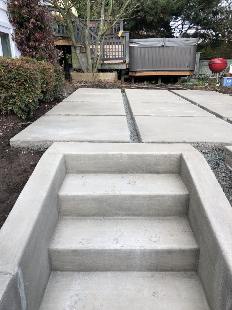 Freshly poured concrete steps leading up to large concrete patio pads in a backyard being landscaped.