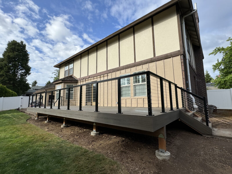 Elevated composite deck with black cable railing wrapping the back of a Tudor-style home under blue skies.