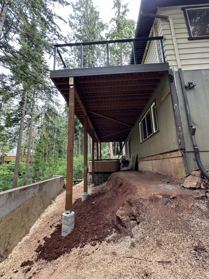 Underside view of an elevated deck on wood posts and concrete footings over a freshly excavated hillside.