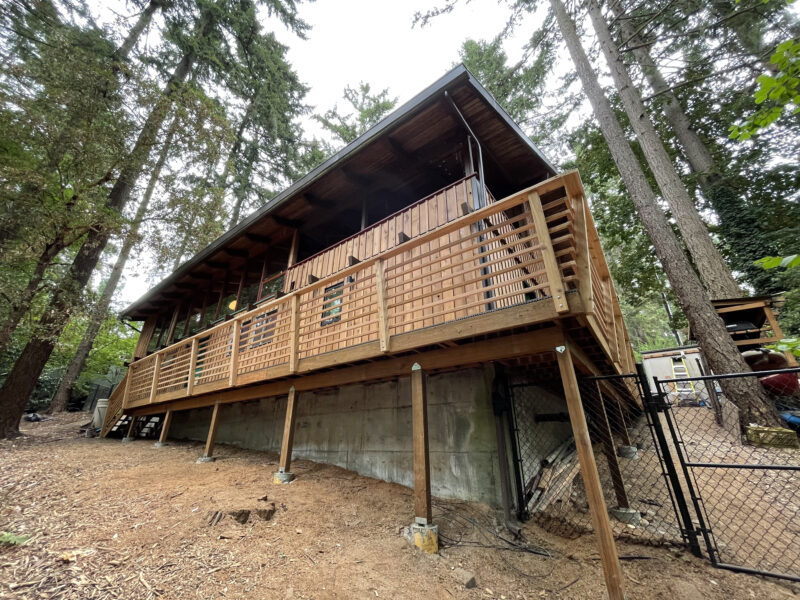 Large elevated cedar deck with lattice-style wood railing wrapping a covered hillside cabin among fir trees.