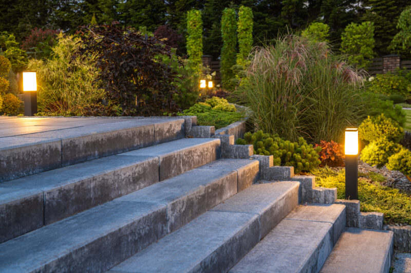 Wide concrete paver stairs with bollard path lights winding through a landscaped garden at dusk.