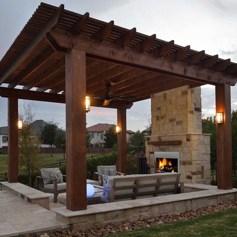 Stained timber pergola with lantern sconces and ceiling fan over a stone outdoor fireplace and lounge seating at dusk.