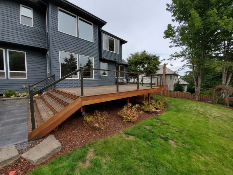 Elevated hardwood deck with cable railing and side stairs on a dark blue two-story home with fresh mulch beds.