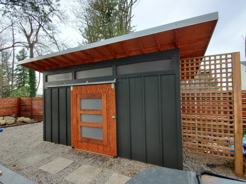 Modern shed with slanted metal roof, dark board-and-batten siding, wood barn door, and cedar lattice privacy screen.