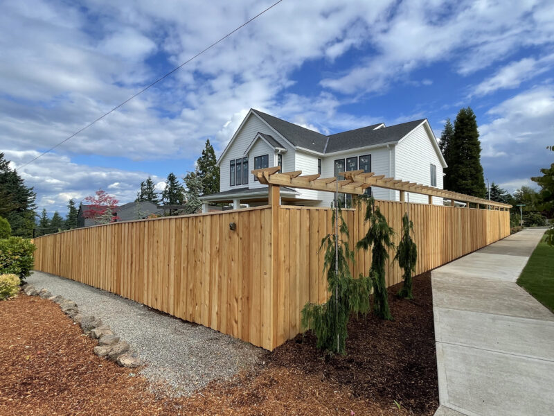 New cedar privacy fence with pergola-style top wrapping a corner lot in front of a farmhouse-style home
