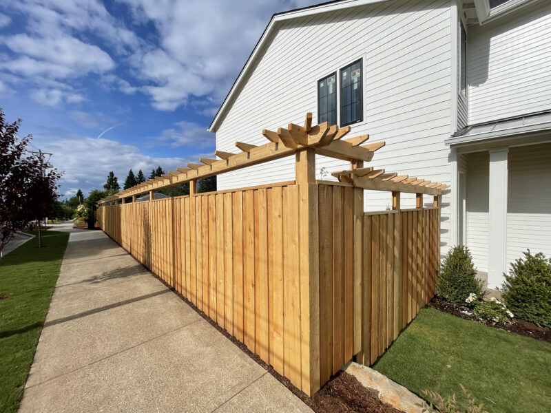 Cedar board fence with decorative pergola-style rafter top running along a sidewalk beside a white house.