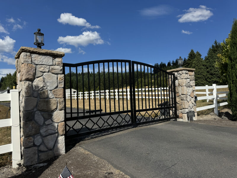 Black wrought-iron driveway entry gate flanked by stone pillar columns with lanterns and white ranch fencing