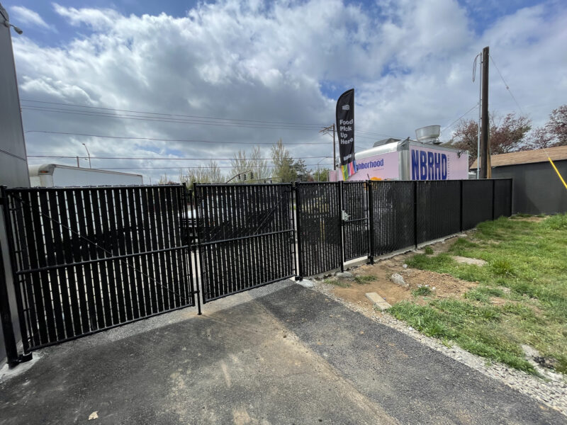 Black chain-link fence with privacy slats and a double swing gate securing a gravel commercial lot.