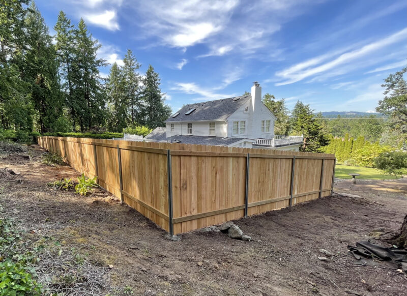 New cedar board fence on steel posts running along a hillside property line with a white farmhouse beyond.